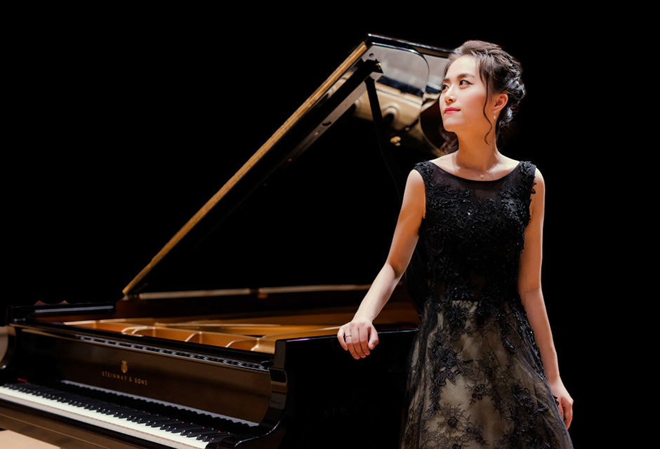 An Asian women, wearing a black dress, standing next to a grand piano, looking into the distance.
